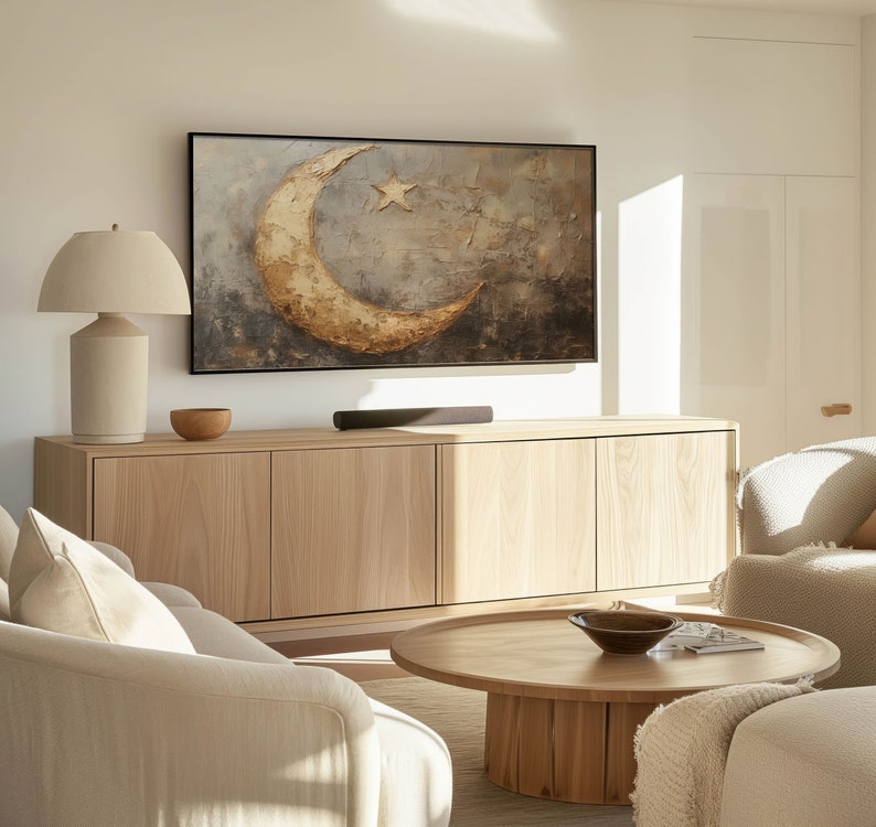 May include: A living room featuring a light wood cabinet, a coffee table, and a sofa. The cabinet has a large flat screen TV on top of it. The TV is displaying a painting of a crescent moon and a star. The coffee table is round and has a wooden top. The sofa is light coloured and has a white throw pillow on it.