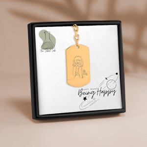 Pode incluir: Um pingente de placa de identificação de cachorro dourado com uma corrente, apresentando um desenho de linha de um cachorro. A placa é exibida em uma caixa preta com o texto "Get Busy Being Happy" e um logotipo para "The Label Lab".