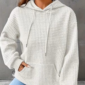 May include: A white, textured hoodie with a waffle-like pattern. The hoodie features a drawstring hood, a front kangaroo pocket, and long sleeves. The fabric appears to be a soft, comfortable material, suitable for casual wear.