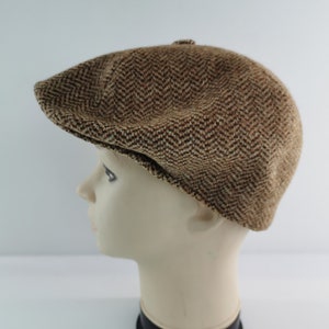 Op de afbeelding: Bruine tweed newsboy cap met een visgraatpatroon. De pet wordt getoond op een mannequinhoofd, waarbij de vorm en textuur worden benadrukt. De pet heeft een afgeronde kroon en een korte, stijve klep.