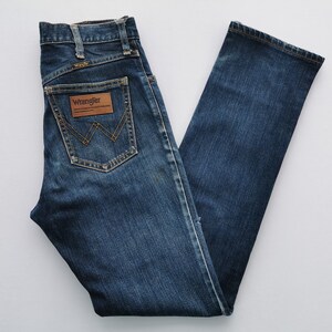 May include: Dark blue denim jeans with the Wrangler logo on the back pocket. The jeans have a classic five-pocket design and a straight leg. The jeans are made of a durable denim material.