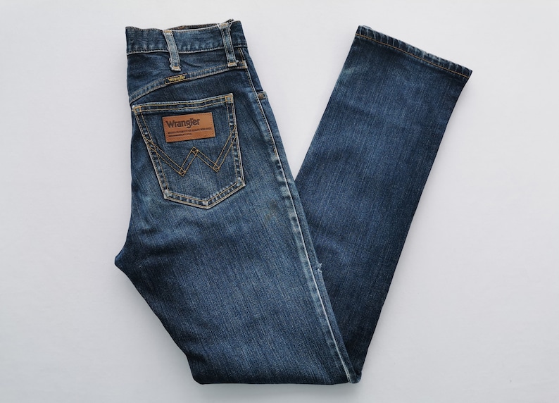 May include: Dark blue denim jeans with the Wrangler logo on the back pocket. The jeans have a classic five-pocket design and a straight leg. The jeans are made of a durable denim material.