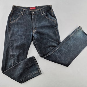 May include: Dark wash denim jeans with a classic five-pocket design. The jeans have a button and zip closure, belt loops, and a straight leg cut. The denim has a slightly faded appearance, with subtle wear marks. The jeans are laid flat on a light grey surface.