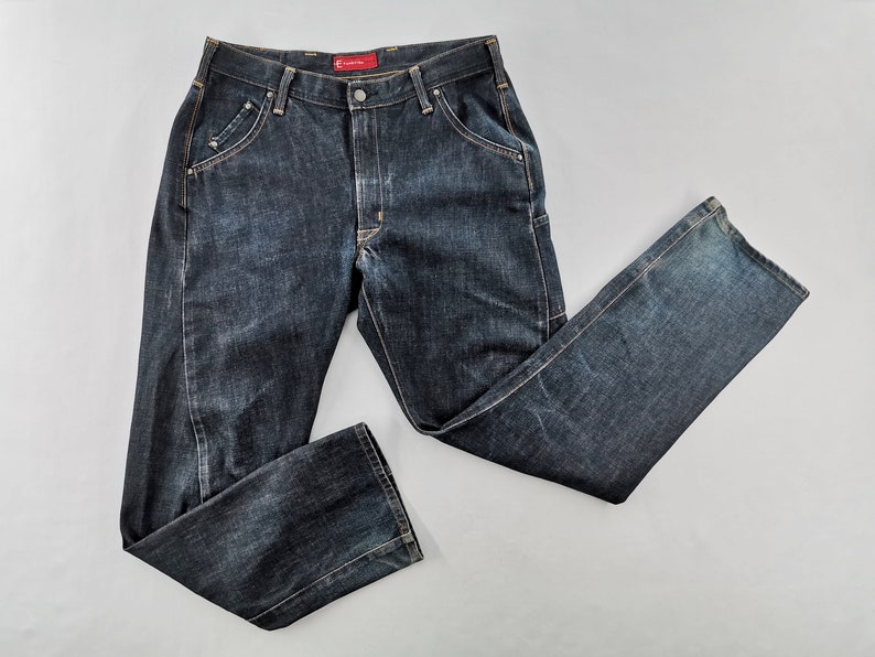 May include: Dark wash denim jeans with a classic five-pocket design. The jeans have a button and zip closure, belt loops, and a straight leg fit. The denim has a slightly faded appearance, with subtle wear marks. The jeans are laid flat on a light grey surface.