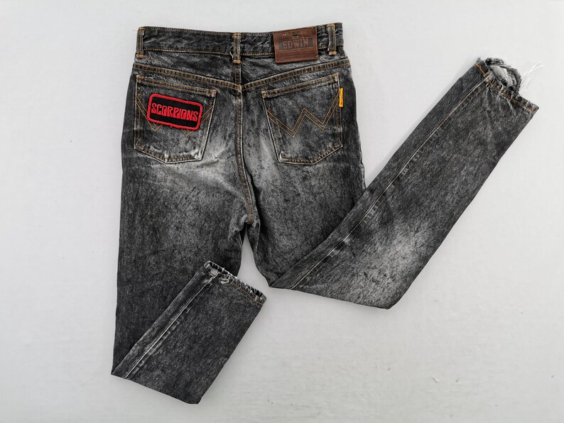 May include: Acid-washed black denim jeans featuring a rectangular red and black Scorpions patch on the left back pocket, a brown leather Edwin brand label on the waistband, and a signature W-stitch design on the right back pocket.
