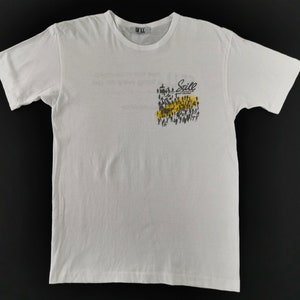 May include: White crew neck t-shirt with a graphic on the front. The graphic features a crowd of people in black and yellow, with the word "Sill" above them. The shirt has a tag that says "Sill".