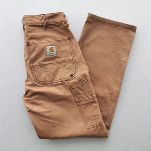 May include: Tan work pants with multiple pockets and a Carhartt logo patch on the back. The pants are made of a durable fabric and have a classic workwear style. The pants are laid flat on a white surface.