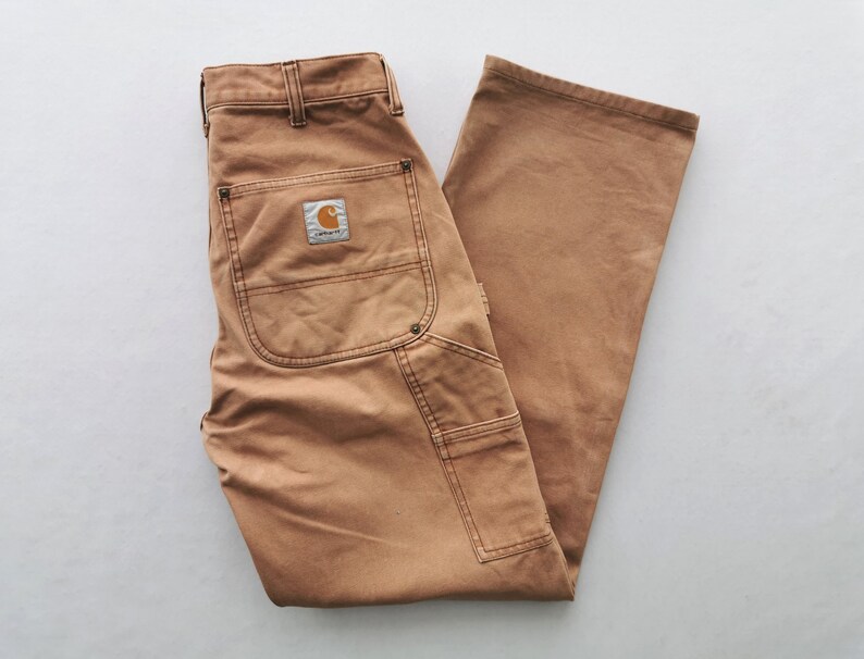May include: Tan work pants with multiple pockets and a Carhartt logo patch on the back. The pants are made of a durable fabric and have a classic workwear style. The pants are laid flat on a white surface.