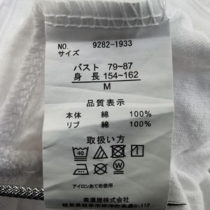 Pu&ograve; includere: Etichetta di tessuto bianca con testo giapponese e simboli di cura. L'etichetta include misure di busto e altezza, e la composizione del tessuto &egrave; 100% cotone. Sono presenti anche istruzioni per il lavaggio.