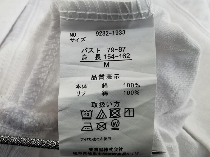 Pu&ograve; includere: Etichetta di tessuto bianca con testo giapponese e simboli di cura. L'etichetta include misure di busto e altezza, e la composizione del tessuto &egrave; 100% cotone. Sono presenti anche istruzioni per il lavaggio.