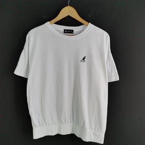 Pu&ograve; includere: T-shirt bianco con girocollo e maniche corte. La maglietta presenta un piccolo logo di canguro nero sul petto. L'orlo inferiore &egrave; arricciato. La maglietta &egrave; appesa a una gruccia di legno su sfondo scuro.
