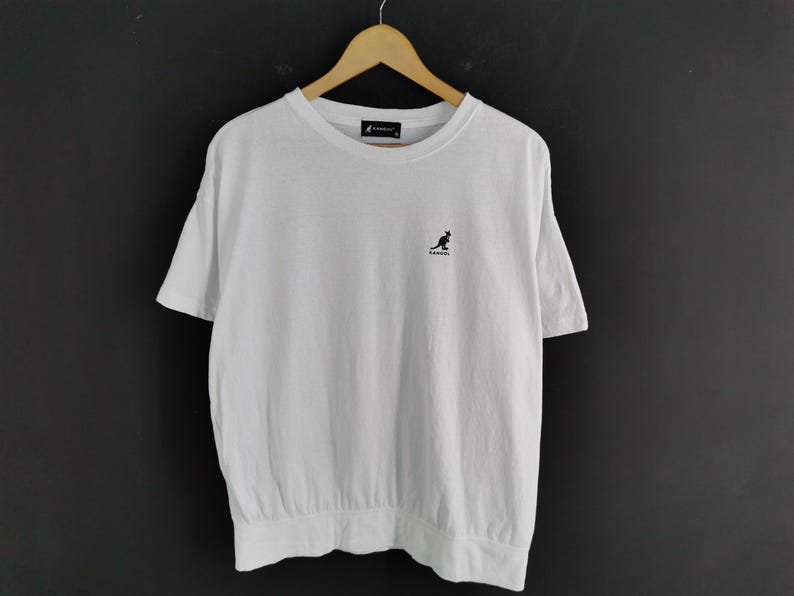 Pu&ograve; includere: T-shirt bianco con girocollo e maniche corte. La maglietta presenta un piccolo logo di canguro nero sul petto. L'orlo inferiore &egrave; arricciato. La maglietta &egrave; appesa a una gruccia di legno su sfondo scuro.