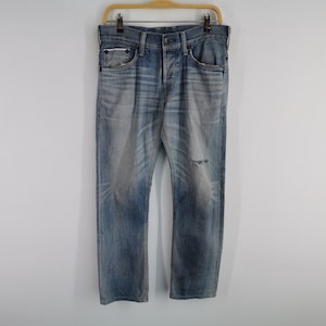 K&ouml;nnte beinhalten: Verblasste blaue Jeans mit einem Used-Look, mit Knopfleiste und einem kleinen Riss am linken Knie. Die Jeans h&auml;ngen an einem Holzb&uuml;gel vor einem schlichten wei&szlig;en Hintergrund.