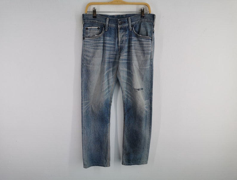 K&ouml;nnte beinhalten: Verblasste blaue Jeans mit einem Used-Look, mit Knopfleiste und einem kleinen Riss am linken Knie. Die Jeans h&auml;ngen an einem Holzb&uuml;gel vor einem schlichten wei&szlig;en Hintergrund.