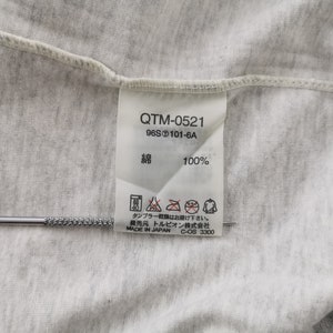 May include: Close-up of a light grey fabric with a tag attached. The tag has the text "QTM-0521", "96S 101-6A", and "綿 100%" printed on it. The tag also includes care instructions and the text "MADE IN JAPAN". A silver tool is placed on the fabric.