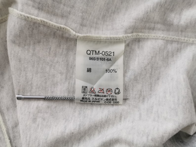 May include: Close-up of a light grey fabric with a tag attached. The tag has the text "QTM-0521", "96S 101-6A", and "綿 100%" printed on it. The tag also includes care instructions and the text "MADE IN JAPAN". A silver tool is placed on the fabric.