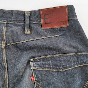 May include: Close-up of the rear of dark wash denim jeans. Features include a brown leather patch with the text "FUNCTION EDWIN", a small red tag, and a pocket with a metal button. The jeans have yellow stitching.