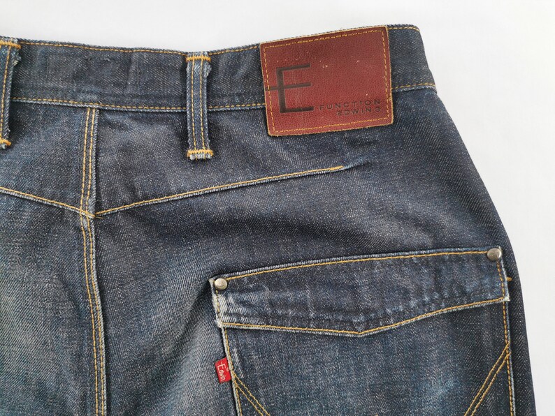 May include: Close-up of the rear of dark wash denim jeans. Features include a brown leather patch with the text "FUNCTION EDWIN", a small red tag, and a pocket with a metal button. The jeans have yellow stitching.