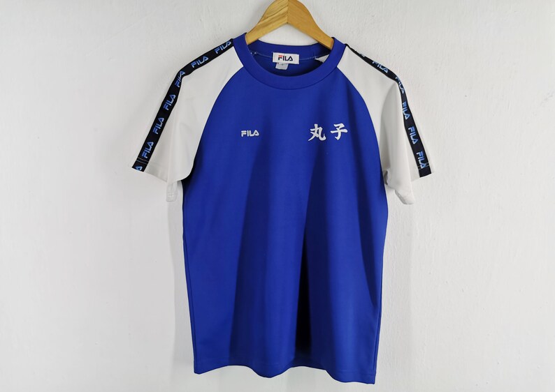 Puede incluir: Camiseta Fila azul y blanca de manga corta. La camiseta tiene mangas blancas y cuerpo azul. El logo de Fila est&aacute; en el pecho y las mangas tienen el logo de Fila impreso. Caracteres japoneses impresos en la parte delantera.