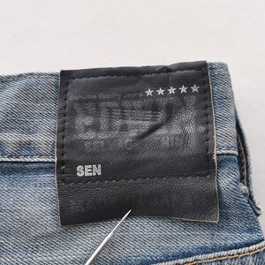 K&ouml;nnte beinhalten: Nahaufnahme einer blauen Jeans mit einem schwarzen Lederaufn&auml;her. Der Aufn&auml;her zeigt gepr&auml;gten Text und f&uuml;nf Sterne. Das Wort "SEN" ist auf dem Aufn&auml;her zu sehen. Die Jeans hat ein klassisches F&uuml;nf-Taschen-Design.