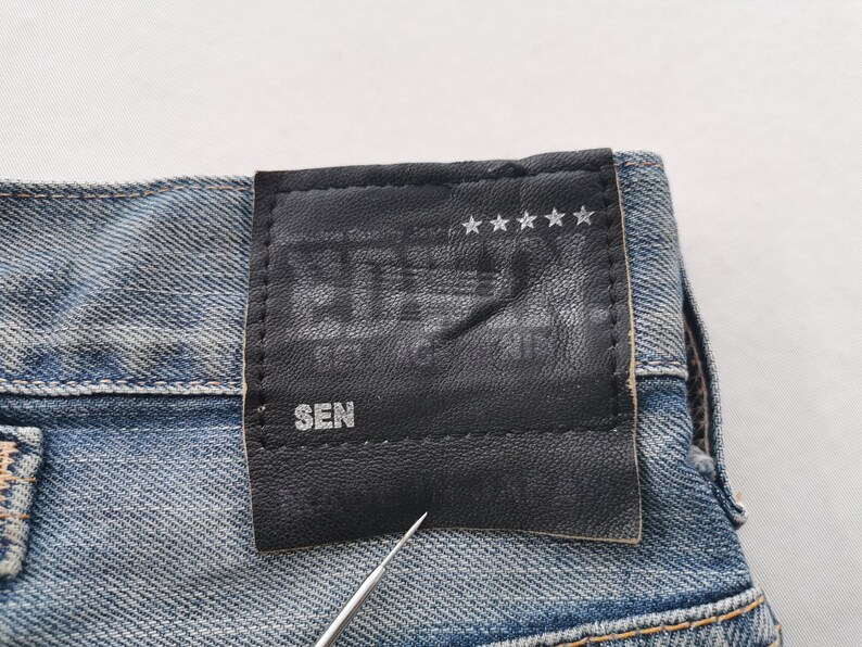 K&ouml;nnte beinhalten: Nahaufnahme einer blauen Jeans mit einem schwarzen Lederaufn&auml;her. Der Aufn&auml;her zeigt gepr&auml;gten Text und f&uuml;nf Sterne. Das Wort "SEN" ist auf dem Aufn&auml;her zu sehen. Die Jeans hat ein klassisches F&uuml;nf-Taschen-Design.