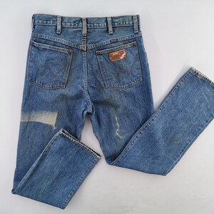 May include: Vintage blue denim jeans with a faded appearance. The jeans feature classic five-pocket styling, a straight leg, and a worn leather patch on the back pocket. The denim shows signs of wear and a light-colored patch.