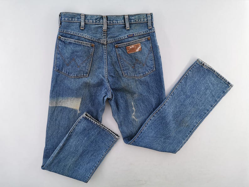 May include: Vintage blue denim jeans with a faded appearance. The jeans feature classic five-pocket styling, a straight leg, and a worn leather patch on the back pocket. The denim shows signs of wear and a light-colored patch.