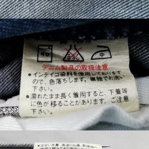 May include: Close-up of a blue denim jeans label with the brand name "Wrangler" in yellow lettering. The label indicates a waist size of 83cm/33. Additional care instructions are printed on a white tag.
