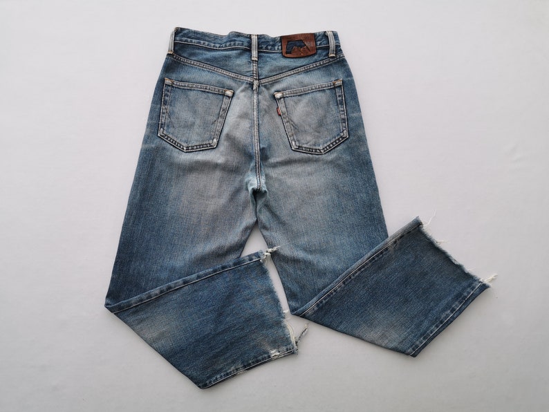 Puede incluir: Vaqueros de mezclilla azul claro con un lavado descolorido y detalles desgastados. Presentan un dise&ntilde;o cl&aacute;sico de cinco bolsillos, un parche de cuero y una etiqueta roja. Los vaqueros tienen una pernera recta con dobladillo sin rematar.