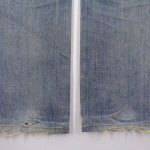 Pu&ograve; includere: Primo piano della parte inferiore di un paio di jeans blu chiaro. I jeans hanno un aspetto usurato con bordi sfilacciati sull'orlo e un piccolo foro vicino al fondo. Il tessuto mostra linee verticali.