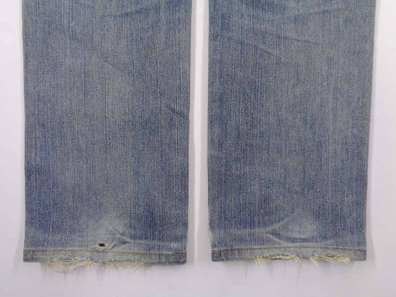 Pu&ograve; includere: Primo piano della parte inferiore di un paio di jeans blu chiaro. I jeans hanno un aspetto usurato con bordi sfilacciati sull'orlo e un piccolo foro vicino al fondo. Il tessuto mostra linee verticali.