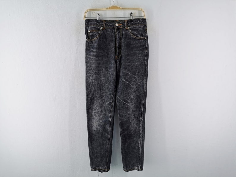 May include: High-waisted jeans in a faded black acid wash finish. These denim pants feature a classic five-pocket design, a metal button closure at the waist, a zip fly, and copper-toned rivets. The fabric displays a mottled, worn-in texture.