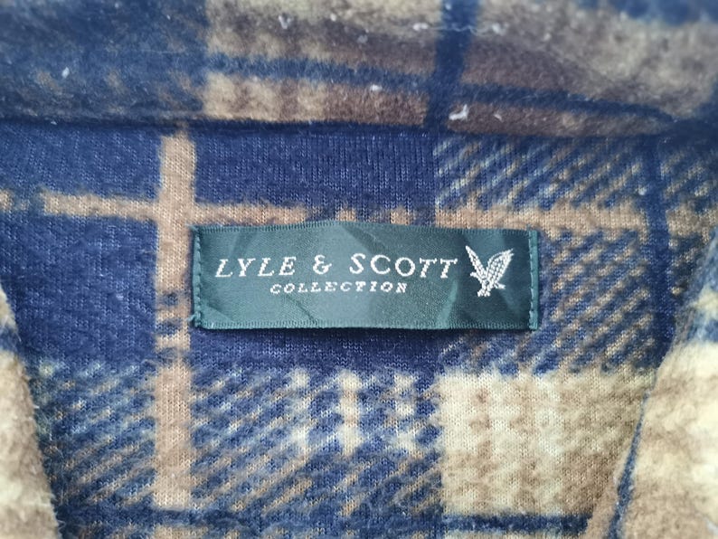 Puede incluir: Primer plano de una tela a cuadros azul, marr&oacute;n y beige. Una etiqueta verde oscuro con el texto "LYLE & SCOTT COLLECTION" y un logotipo de &aacute;guila blanca est&aacute; cosida a la tela.