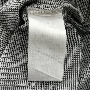May include: A white rectangular garment care label attached to a black and white textured fabric. The label features faint, partially obscured text and small laundry instruction symbols printed in a light gray ink at the top of the tag.
