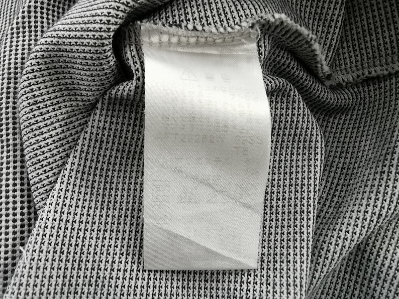 May include: A white rectangular garment care label attached to a black and white textured fabric. The label features faint, partially obscured text and small laundry instruction symbols printed in a light gray ink at the top of the tag.