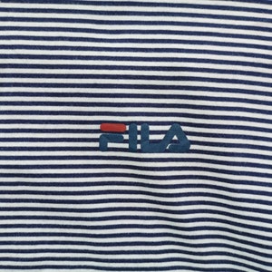 Puede incluir: Primer plano de una camiseta a rayas azul marino y blanca. La prenda presenta el logotipo "FILA" en azul marino con un acento rojo. Las rayas son horizontales y est&aacute;n espaciadas uniformemente.