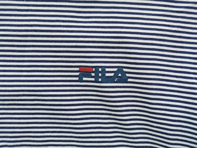 Puede incluir: Primer plano de una camiseta a rayas azul marino y blanca. La prenda presenta el logotipo "FILA" en azul marino con un acento rojo. Las rayas son horizontales y est&aacute;n espaciadas uniformemente.