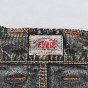 May include: Close-up of a section of dark gray denim jeans. The jeans have orange stitching and a rectangular label that reads "EDWIN Genuine Quality Jeans" in red and white. The label also includes the text "Exclusive design for lasting goodlook."