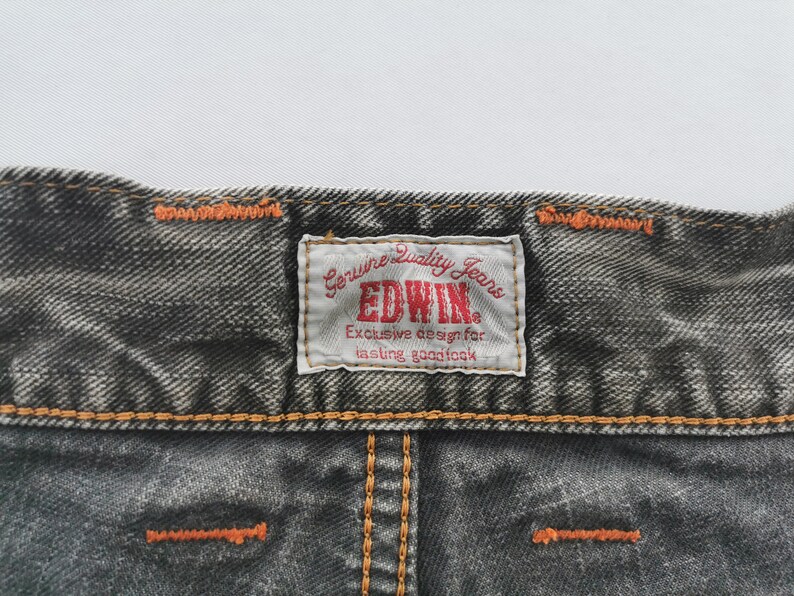May include: Close-up of a section of dark gray denim jeans. The jeans have orange stitching and a rectangular label that reads "EDWIN Genuine Quality Jeans" in red and white. The label also includes the text "Exclusive design for lasting goodlook."