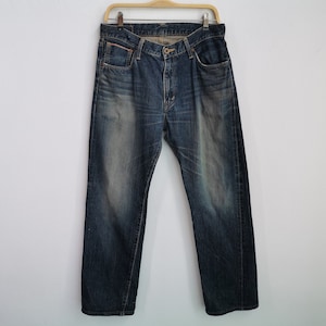 Pu&ograve; includere: Jeans di denim blu scuro appesi a una gruccia di legno. I jeans hanno un classico design a cinque tasche, una chiusura con bottone e cerniera e un aspetto sbiadito. I jeans sono di peso medio e sembrano pre-indossati.