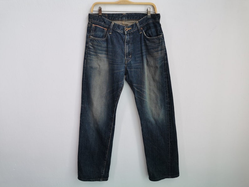 Pu&ograve; includere: Jeans di denim blu scuro appesi a una gruccia di legno. I jeans hanno un classico design a cinque tasche, una chiusura con bottone e cerniera e un aspetto sbiadito. I jeans sono di peso medio e sembrano pre-indossati.