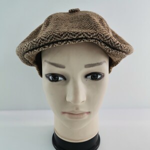 Op de afbeelding: Een bruine tweed newsboy cap met een afgeronde kroon en een kleine, stijve klep. De pet wordt gepresenteerd op een mannequinhoofd tegen een witte achtergrond. De pet heeft een textuur.