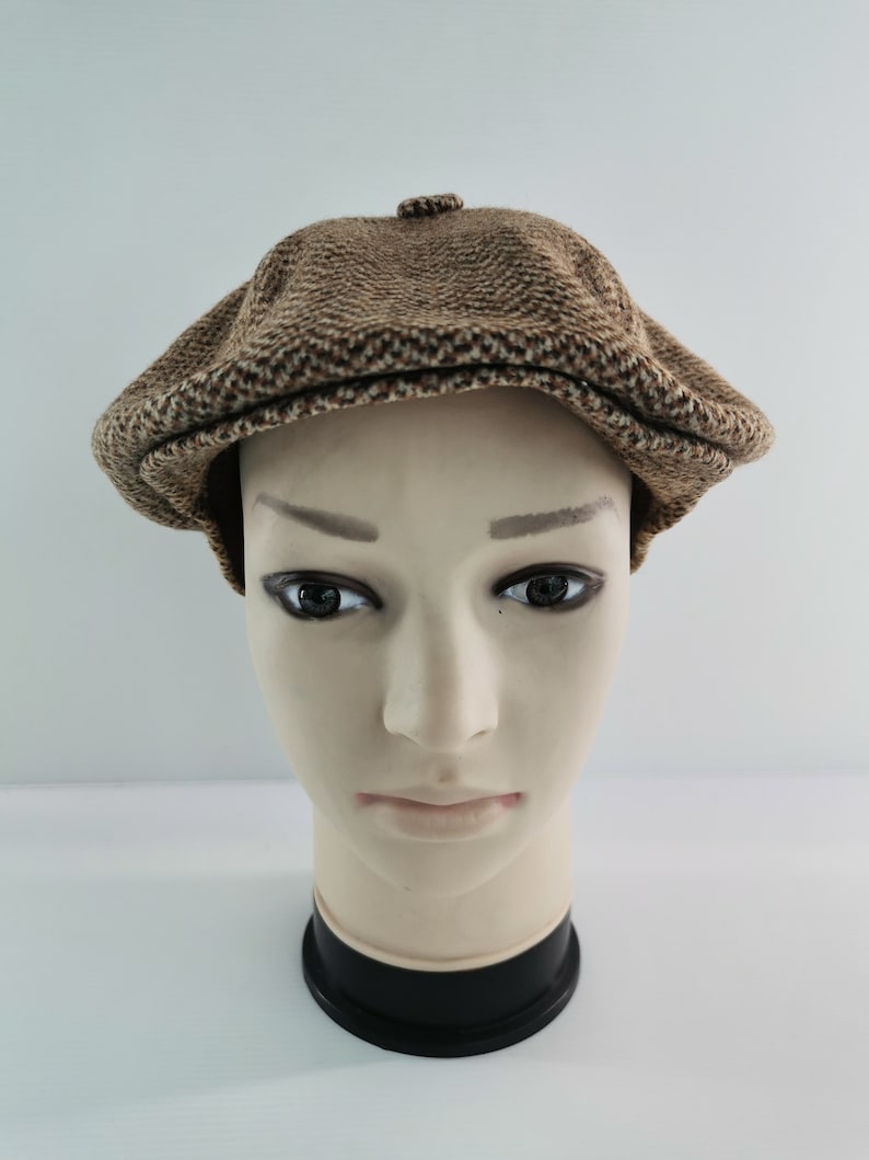 Op de afbeelding: Een bruine tweed newsboy cap met een afgeronde kroon en een kleine, stijve klep. De pet wordt gepresenteerd op een mannequinhoofd tegen een witte achtergrond. De pet heeft een textuur.