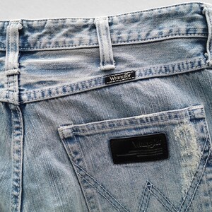 Pu&ograve; includere: Primo piano di jeans di denim blu chiaro con il logo Wrangler sulla tasca posteriore e sulla cintura. I jeans hanno un aspetto usurato con aree sbiadite e cuciture visibili.
