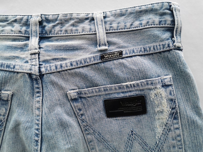 Pu&ograve; includere: Primo piano di jeans di denim blu chiaro con il logo Wrangler sulla tasca posteriore e sulla cintura. I jeans hanno un aspetto usurato con aree sbiadite e cuciture visibili.