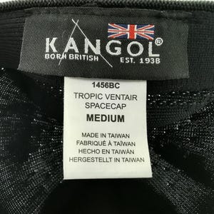 Puede incluir: Primer plano de un sombrero Kangol Tropic Ventair Spacecap negro. El sombrero presenta una etiqueta con el logotipo de Kangol, la bandera brit&aacute;nica y el texto "BORN BRITISH EST. 1938". La etiqueta tambi&eacute;n incluye el estilo 1456BC y la talla "MEDIUM".