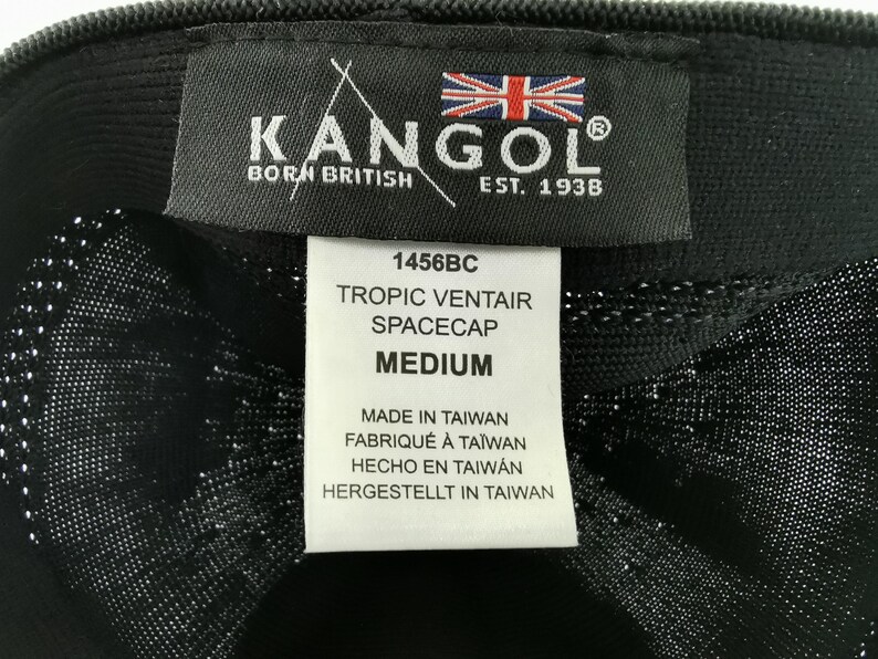 Puede incluir: Primer plano de un sombrero Kangol Tropic Ventair Spacecap negro. El sombrero presenta una etiqueta con el logotipo de Kangol, la bandera brit&aacute;nica y el texto "BORN BRITISH EST. 1938". La etiqueta tambi&eacute;n incluye el estilo 1456BC y la talla "MEDIUM".