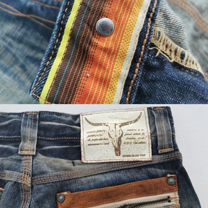 Pu&ograve; includere: Primo piano di jeans in denim con un design dettagliato. I jeans presentano una cintura a righe colorate, una toppa in pelle con un teschio di toro e una tasca con cerniera e tiretto in pelle. I jeans sono blu sbiadito.