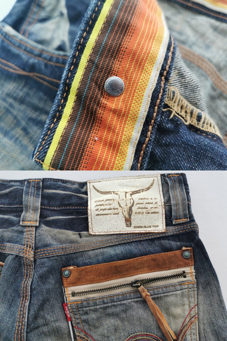 Pu&ograve; includere: Primo piano di jeans in denim con un design dettagliato. I jeans presentano una cintura a righe colorate, una toppa in pelle con un teschio di toro e una tasca con cerniera e tiretto in pelle. I jeans sono blu sbiadito.