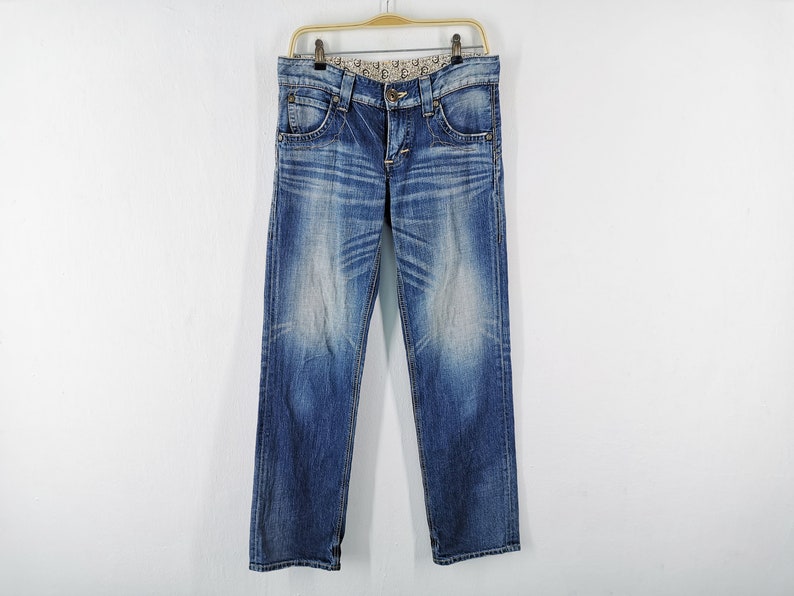May include: Blue denim jeans with a faded wash effect, hanging on a gold-colored hanger. The jeans have a classic five-pocket design, a button and zipper closure, and a decorative pattern on the waistband.